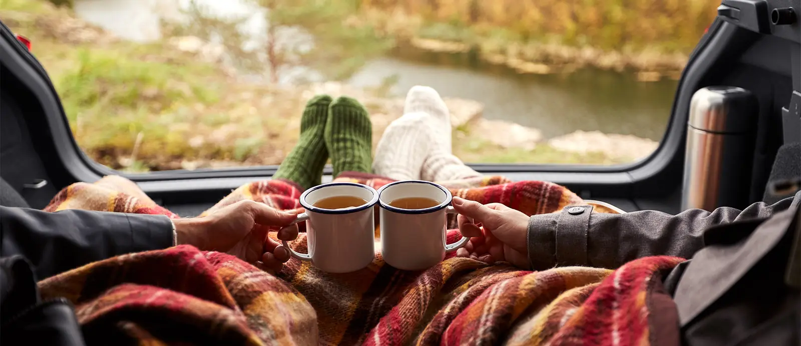 camping couple sitting in car under blanket and toasting tea cups